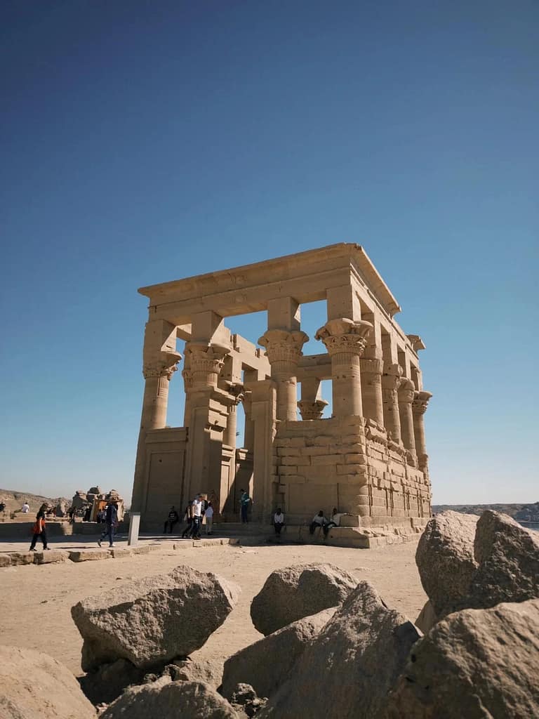 Visitors walk around and rest near the grand stone columns of the Philae Temple in Aswan, Egypt, under a clear blue sky. The well-preserved ancient structure features intricately carved capitals and massive stone blocks. Large boulders in the foreground add depth and frame the historic temple, which stands as a striking symbol of ancient Egyptian architecture.