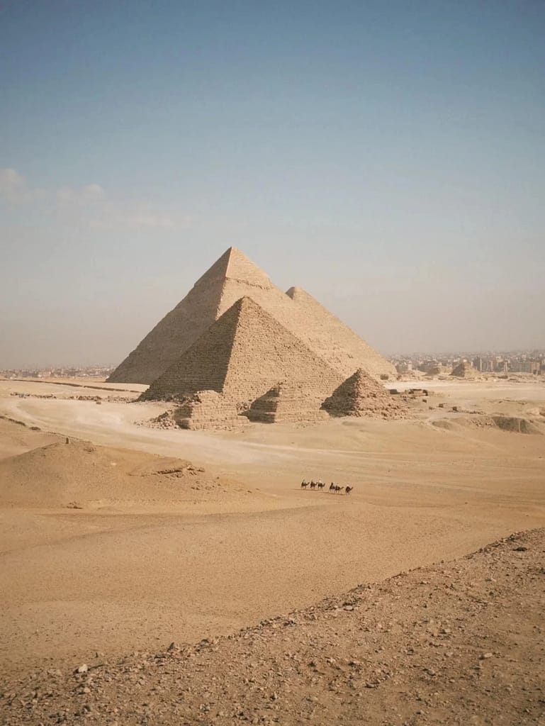 Wide-angle view of the Pyramids of Giza in Egypt under a clear blue sky, showcasing the Great Pyramid of Khufu, the Pyramid of Khafre, and the smaller Pyramid of Menkaure. The foreground features a vast expanse of golden desert with a small group of camels and riders traversing the sandy terrain. The distant cityscape of Cairo is faintly visible on the horizon.
Egypt solar Eclipse tours