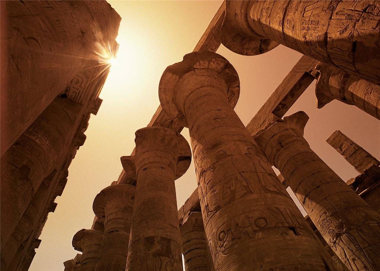 Low-angle view of massive sandstone columns at the Karnak Temple in Luxor, Egypt, with intricate hieroglyphs and carvings etched into their surfaces. The sun shines dramatically through a gap between the towering pillars, casting a warm golden light and creating a powerful sense of scale and ancient grandeur.