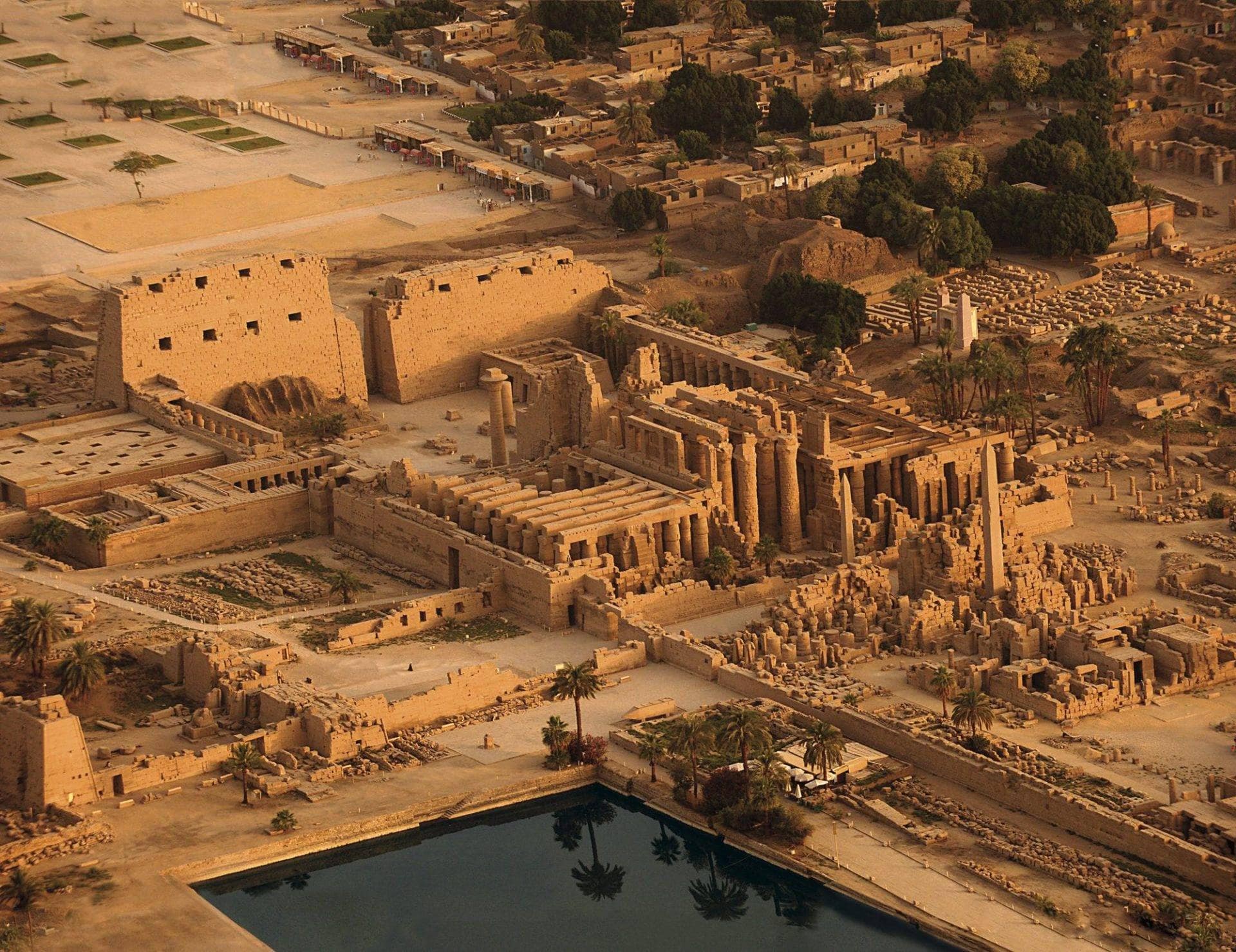 Aerial view of the Karnak Temple Complex in Luxor, Egypt, showcasing its massive ancient sandstone structures, including tall columns, pylons, and obelisks. The scene is bathed in warm, golden light, highlighting the temple’s architectural grandeur. In the foreground, a sacred rectangular water basin reflects the temple. Surrounding the complex are clusters of palm trees and the modern city.
